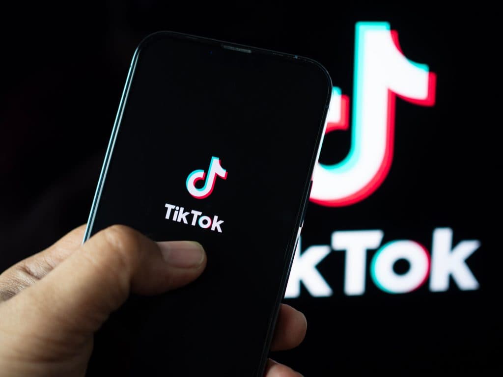 Na imagem, uma mão segura um celular cuja tela apresenta o logo do TikTok