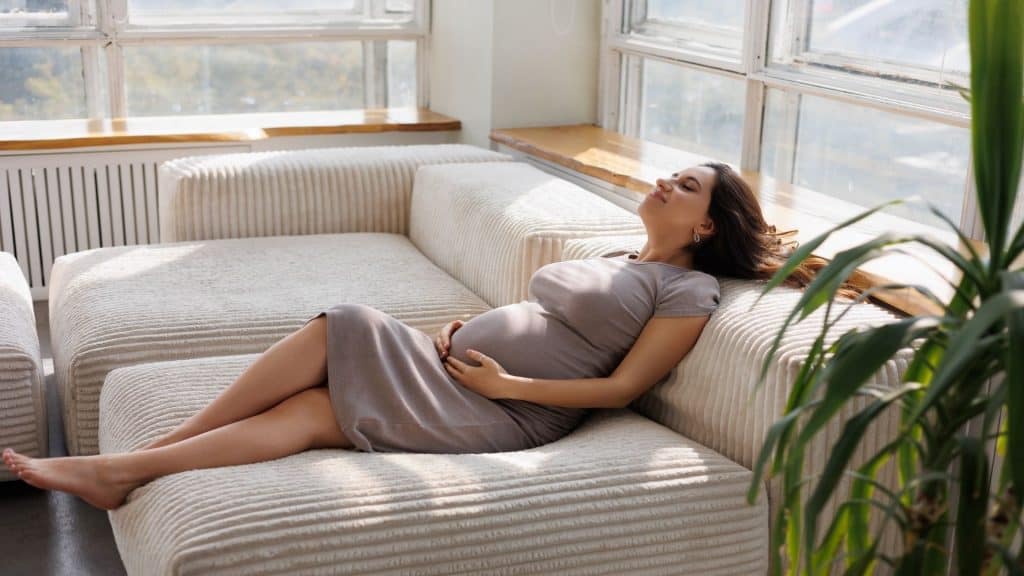 Linda mulher grávida em vestido bege descalça. Barriga de grávida bem grande. Bonita mulher morena esperando um filho. Senhora em seu quarto deitada no sofá perto da janela.