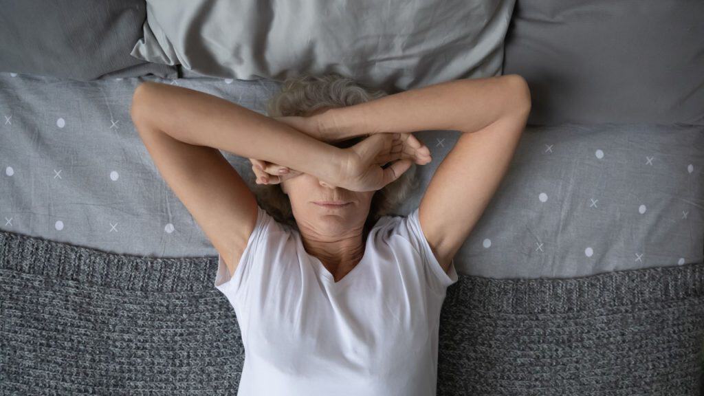 Mulher idosa pacífica dormindo de manhã ou à tarde, descansando na cama, cobrindo os olhos com as mãos da luz do dia. Senhora de idade deitada no quarto, sofrendo de dor de cabeça, enxaqueca, insônia. Vista de cima.