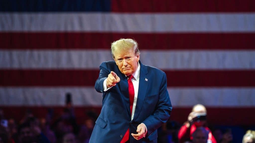 Donald Trump apontando para a frente; atrás, parte da bandeira dos EUA desfocada