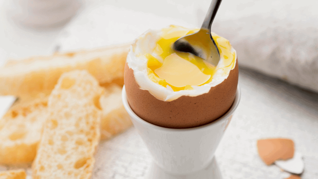 Ovo cozido com gema mole servido em suporte branco, com uma colher mergulhada na gema e fatias de pão ao fundo