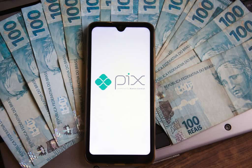 Logo do Pix na tela de um smartphone; debaixo dele, várias notas de R$ 100