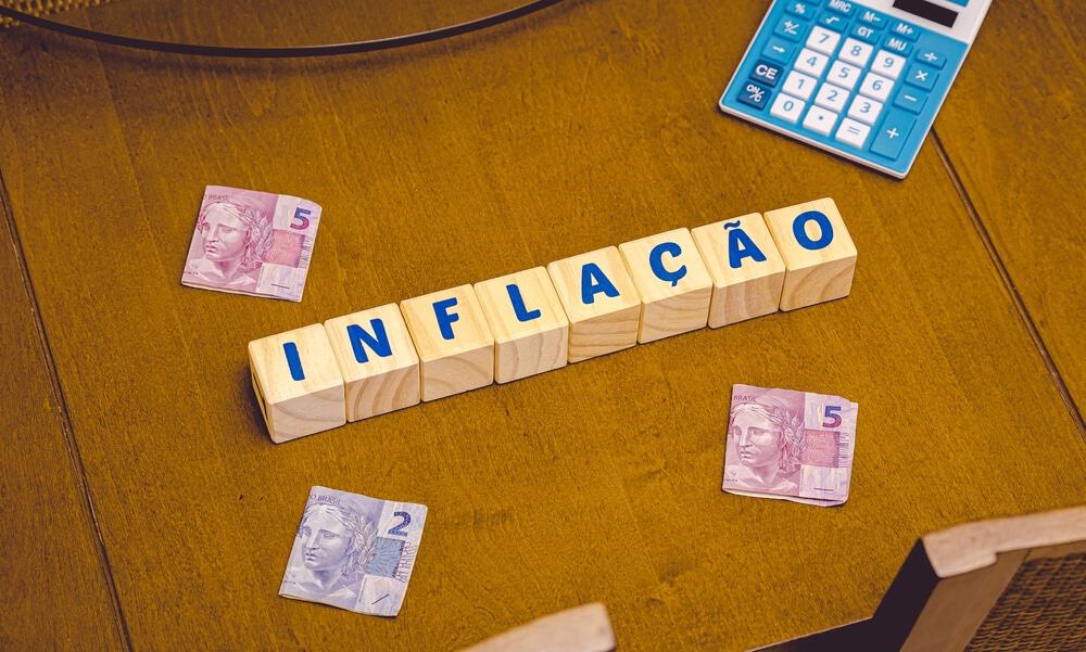 imagem destaca blocos de madeira com letras que formam a palavra INFLAÇÃO. tudo está em cima de uma mesa, onde também há notas de cinco reais