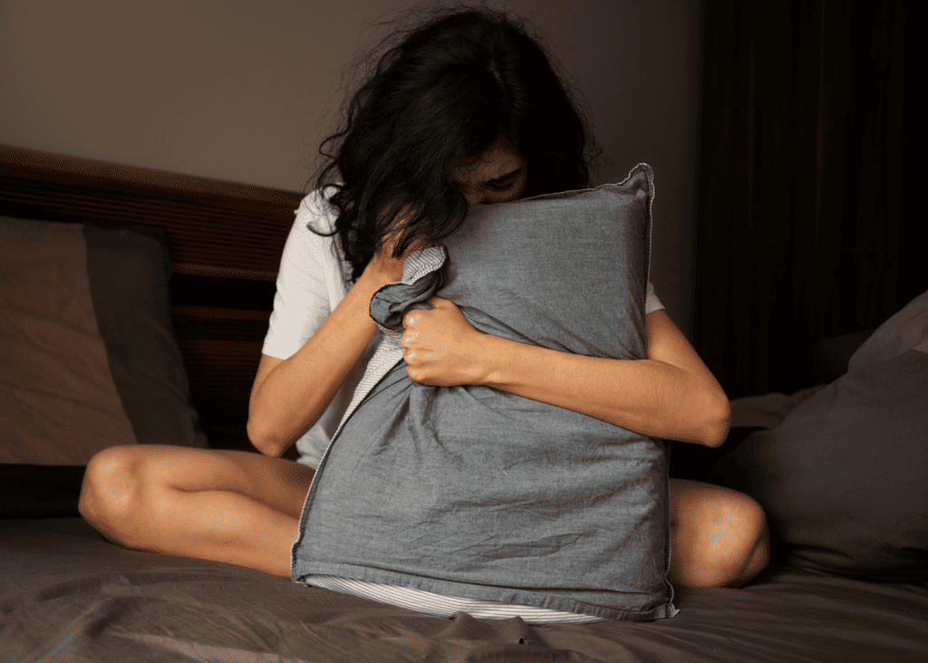 imagem mostra uma mulher sentada na cama, triste, com o rosto afundado em um travesseiro