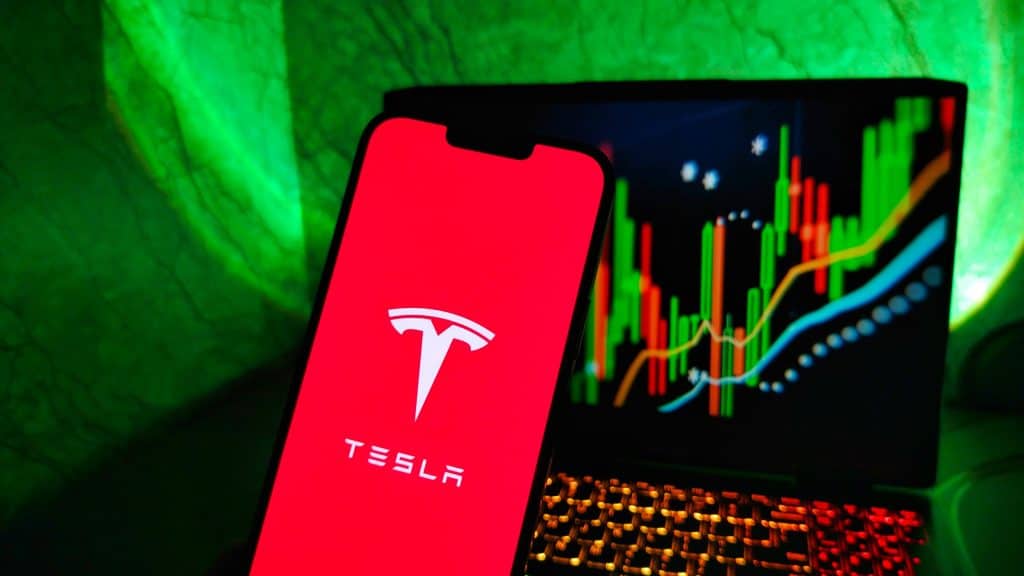 À frente, logo da Tesla em um smartphone; ao fundo, um notebook com gráficos