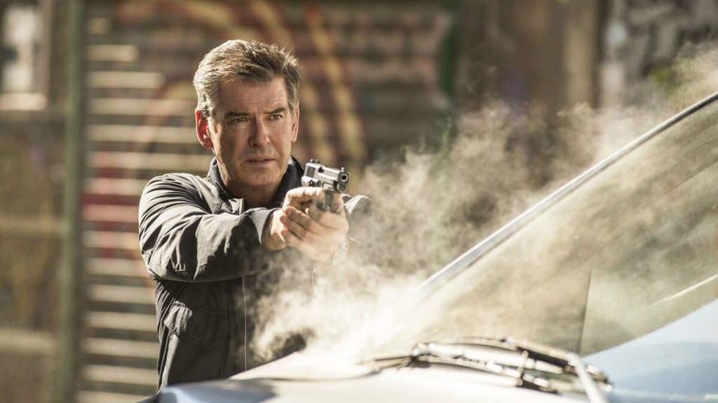 imagem mostra um homem adulto grisalho apontando um revólver sobre o capô enfumaçado de um carro