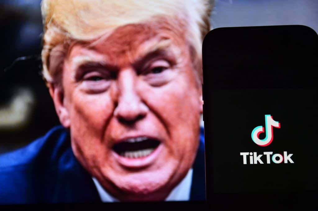 Ao fundo, meio desfocado, rosto de Donald Trump com a boca aberta; à frente, logo do TikTok em um smartphone
