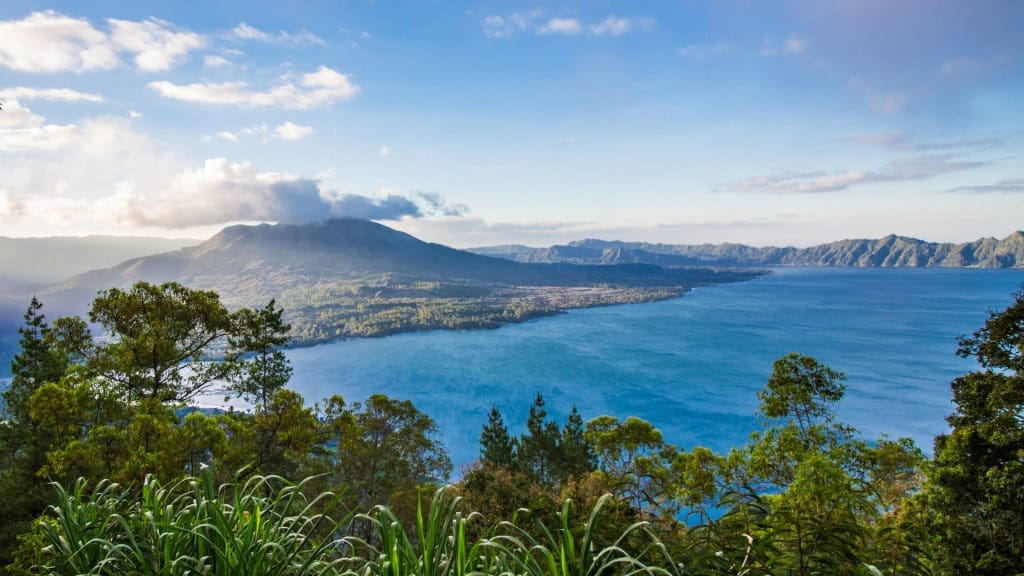 🌋 Em primeiro plano, um grande lago azul reflete a luz suave do céu. À sua margem, há vegetação verdejante e pequenas edificações. Ao fundo, destaca-se um imponente vulcão parcialmente envolto por nuvens, criando uma atmosfera majestosa e misteriosa na paisagem.
