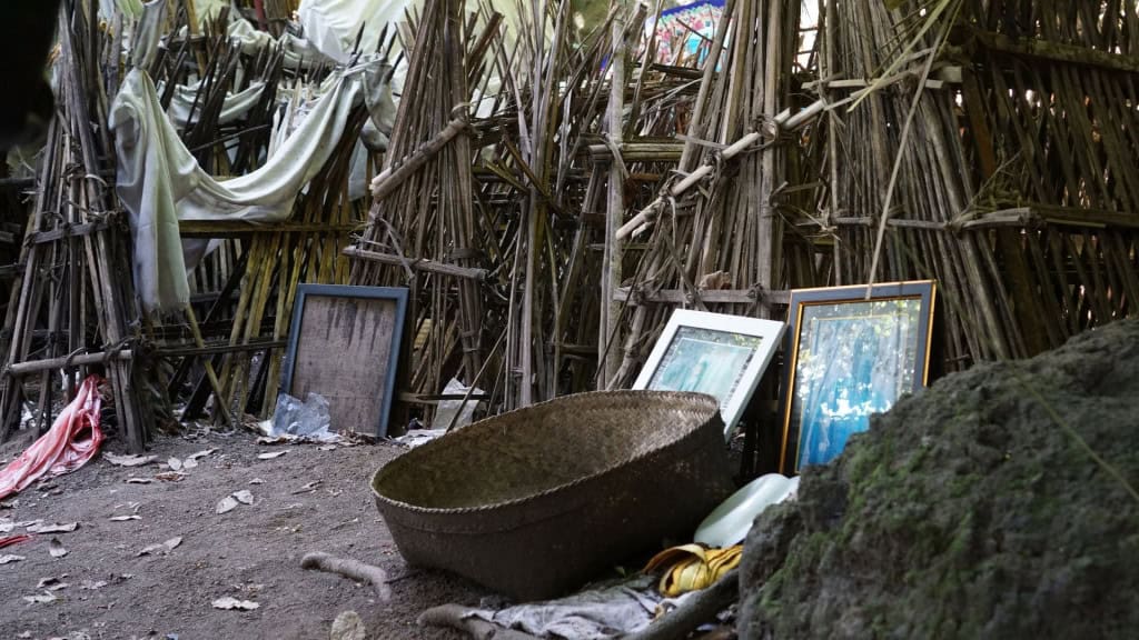 A imagem mostra estruturas feitas com galhos e gravetos amarrados com cordas, algumas cobertas por tecido branco. Em frente a elas há fotos emolduradas encostadas, um cesto de palha grande e objetos como um recipiente plástico branco e um pano amarelo, sobre um chão de terra com folhas secas.