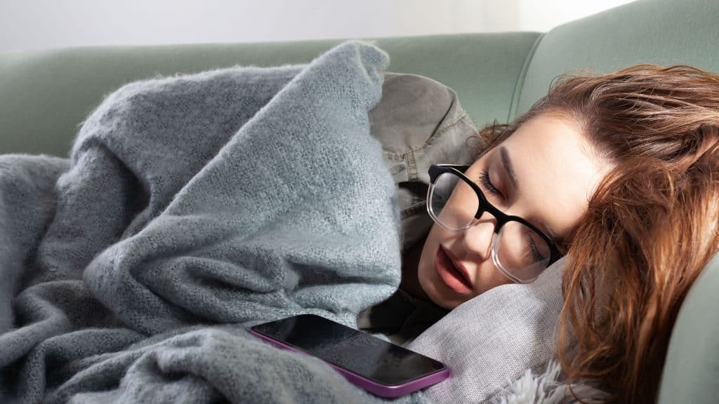 Jovem mulher exausta do vício do smartphone, dorme no sofá com seu telefone. Conceito de excesso de navegação nas redes sociais, uso excessivo da Internet e conexão on-line constante