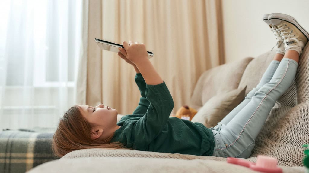 Menina pequena e fofa jogando em um tablet deitada no sofá com os pés para o alto e tênis calçados, sozinha em casa em uma sala grande. Crianças e tecnologias modernas