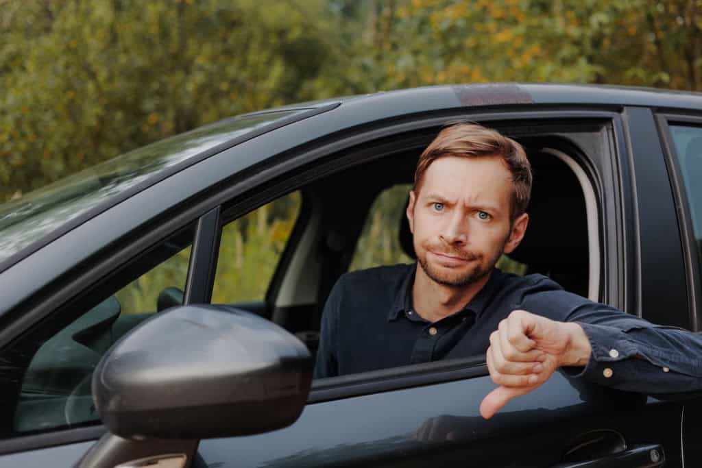 Imagem mostra motorista homem descontente dentro do carro fazendo sinal com gesto que indica problemas com polegar apontado para baixo