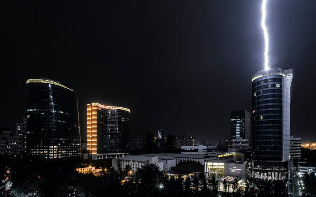 imagem mostra uma cidade à noite, no escuro. Na foto, uma raja de raio cai do céu e é puxada até o para-raio no terraço de um grande edifício