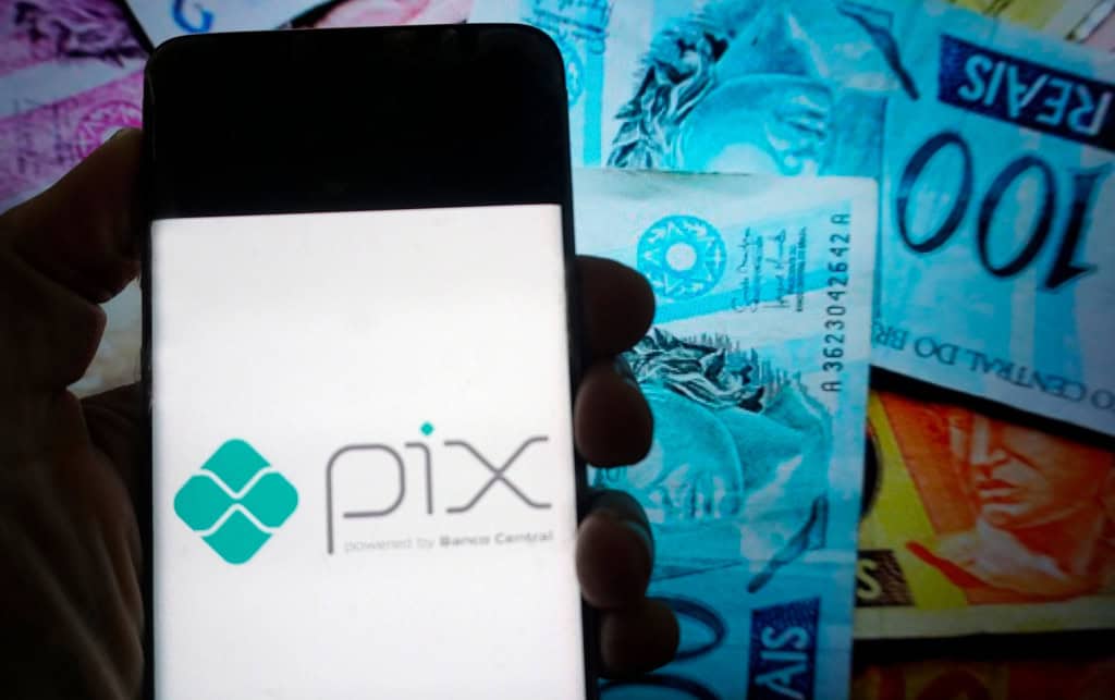 Ao fundo, notas de real; à frente, o logotipo do Pix em um smartphone