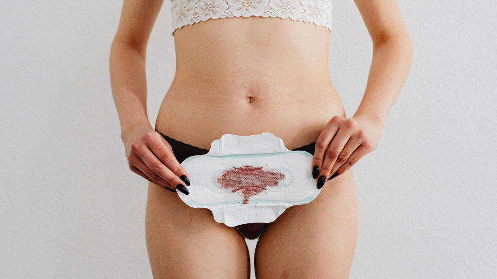 A imagem mostra uma pessoa de calcinha preta e top branco rendado segurando um absorvente com sangue menstrual. A cena retrata um momento íntimo relacionado ao ciclo menstrual