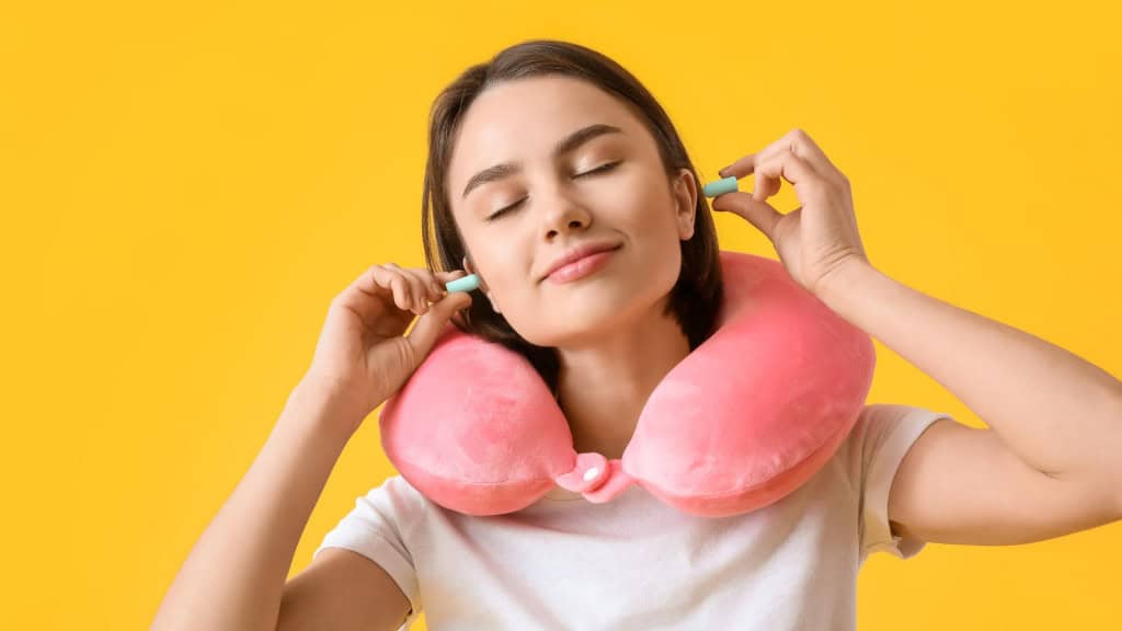 "Jovem mulher com travesseiro de pescoço colocando protetores auriculares sobre fundo amarelo."