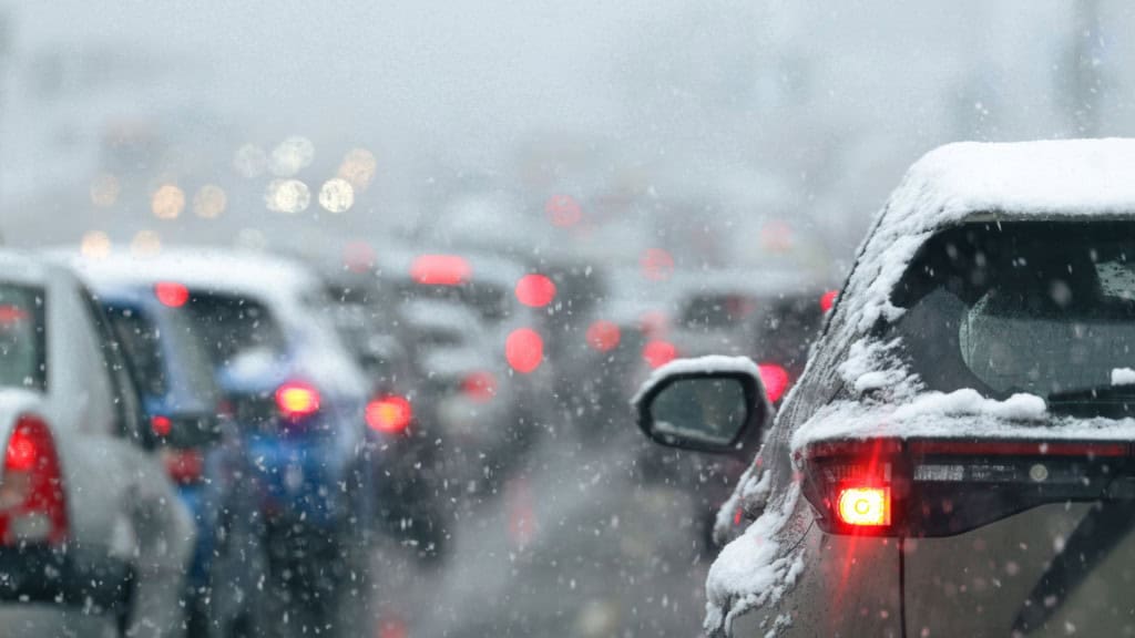 Uma estrada coberta de neve mostra um longo congestionamento, com carros parados e faróis acesos em meio à nevasca. A neve intensa e a baixa visibilidade indicam condições perigosas, exigindo atenção redobrada dos motoristas. O cenário revela os desafios do trânsito urbano em dias de tempestade de inverno.