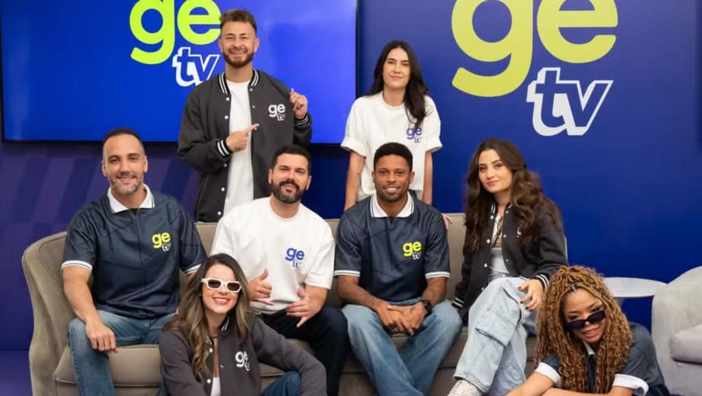 imagem mostra uma equipe de profissionais de comunicação: alguns em pé e outros sentados num sofá. no fundo, há a imagem/logo do program GE TV