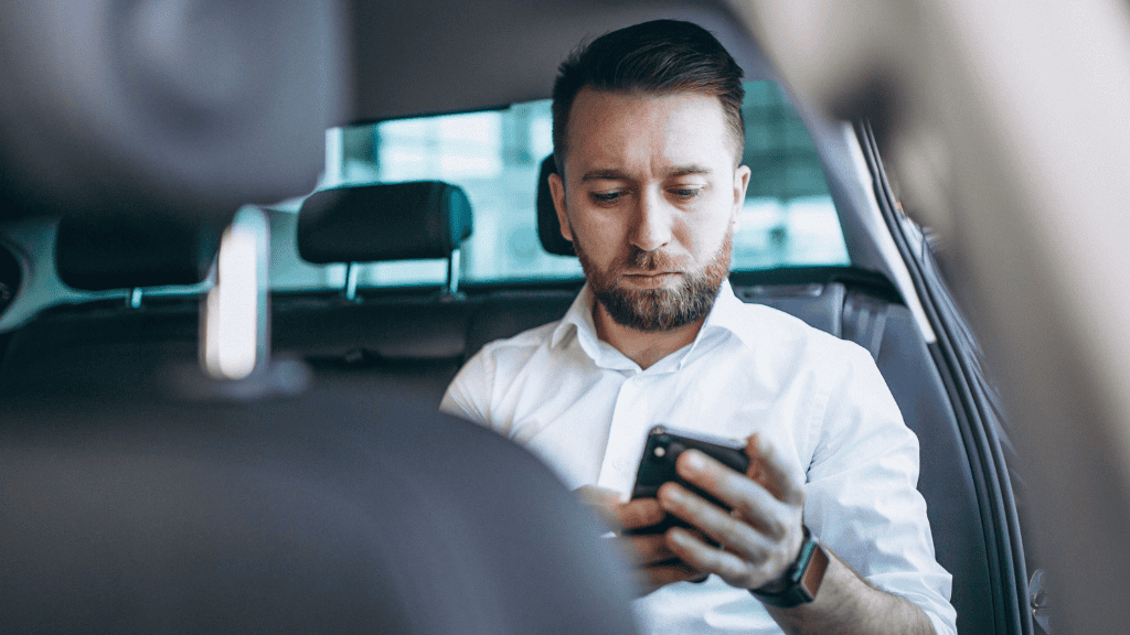 Homem sério sentado no banco traseiro de um Uber, olhando o celular com expressão concentrada, visto do ponto de vista do banco da frente