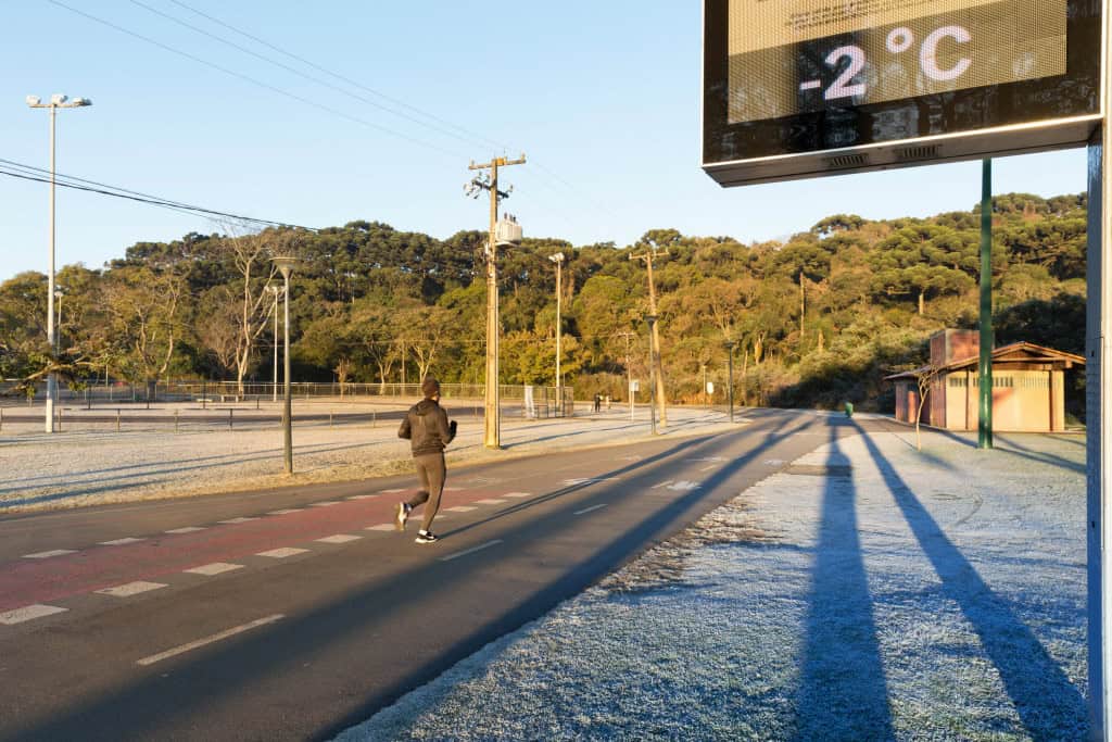 Estrada ensolarada com neve na lateral direita da foto; acima, um termômetro de rua marcando −2 °C