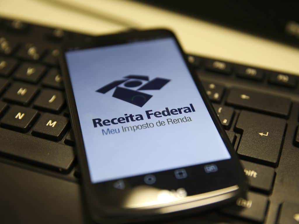 Celular com homepage da Receita Federal aberta em navegador; aparelho está sobre teclado de computador