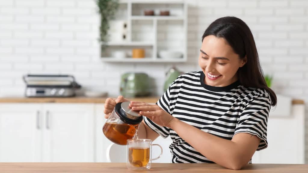Linda jovem mulher feliz derramando chá quente de chaleira transparente em xícara na cozinha