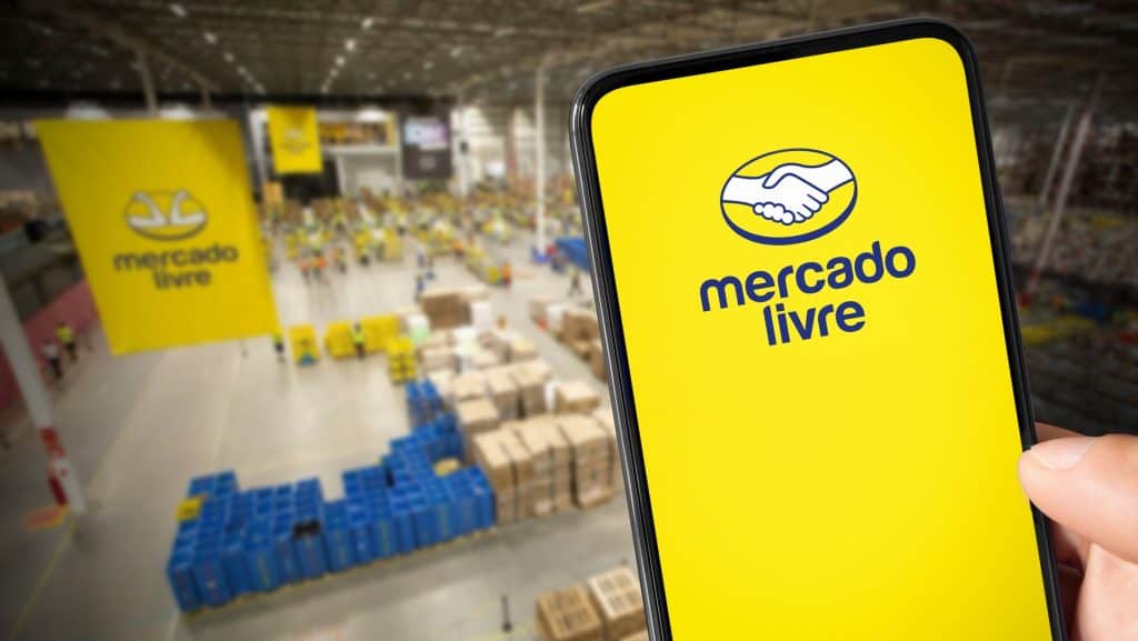 Pessoa segurando celular com logomarca do Mercado Livre na tela; no fundo, um armazém da varejista