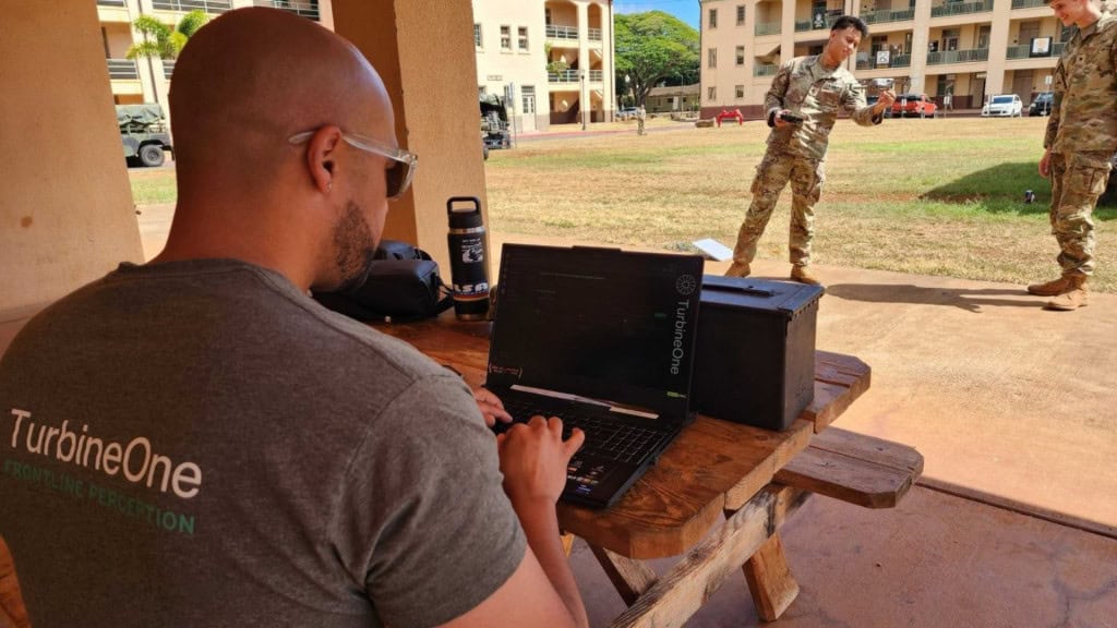 Homem com camiseta da startup de inteligência artificial (IA) TurbineOne usando notebook enquanto soldados do Exército dos EUA preparam drone