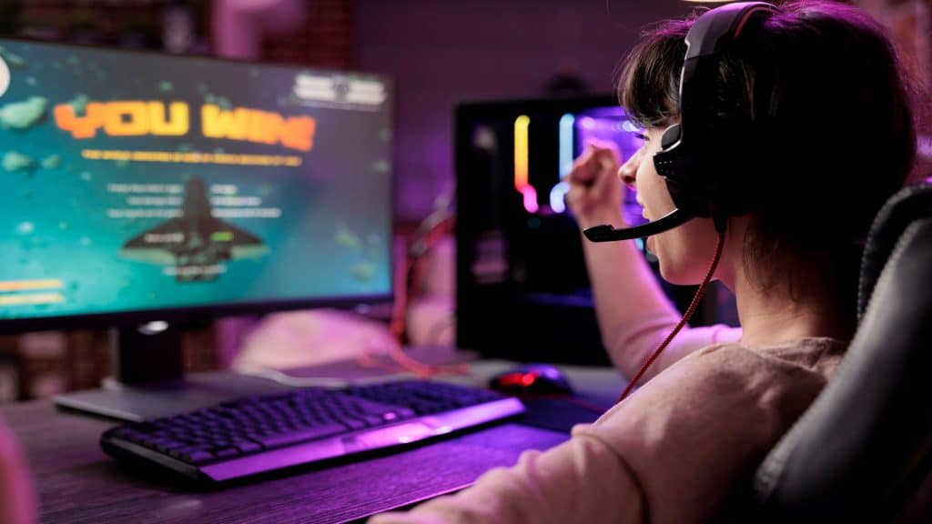 Jogadora empolgada comemorando a vitória em um duelo de videogame multiplayer online de naves espaciais, derrotando os oponentes. Mulher aproveitando seu tempo livre em um apartamento com luzes RGB, se sentindo nas nuvens com a vitória.